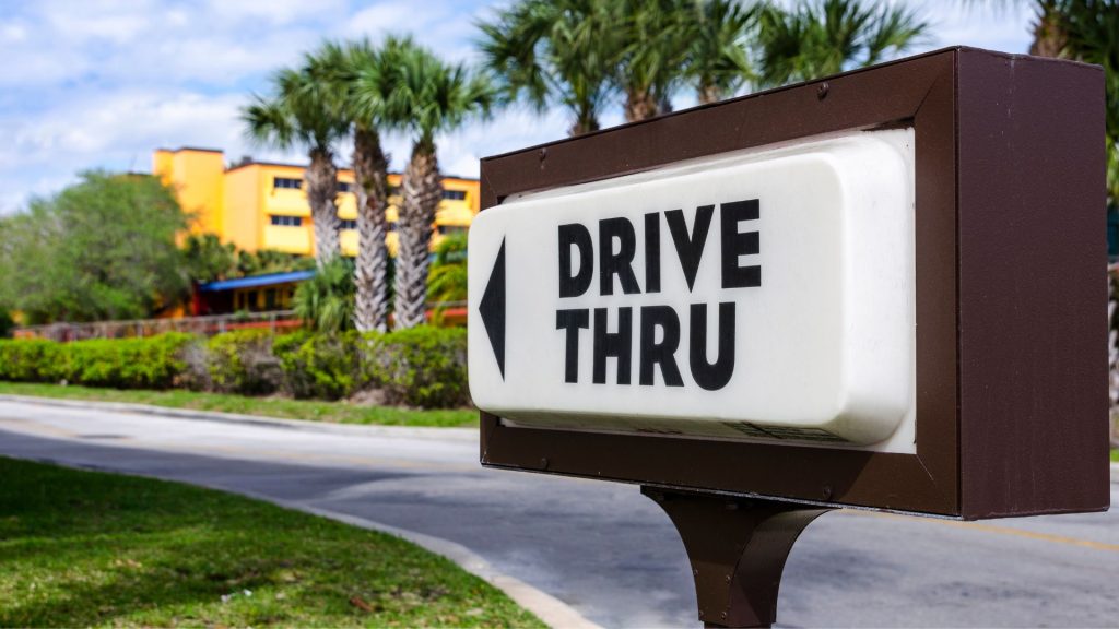 This is a photograph of a drive-thru sign in front of a blurred background featuring a sunny, landscaped area with palm trees and a brightly colored building. The sign is prominently placed in the foreground and has a dark frame, with a backlit section that reads "DRIVE THRU" in bold, capitalized letters, and an arrow indicating the direction to the drive-thru lane. The setting appears to be tropical or subtropical, suggested by the presence of palm trees and the bright, warm colors of the building. The focus on the sign with the background softly blurred indicates the primary importance of the drive-thru service being advertised, and may suggest convenience and accessibility.