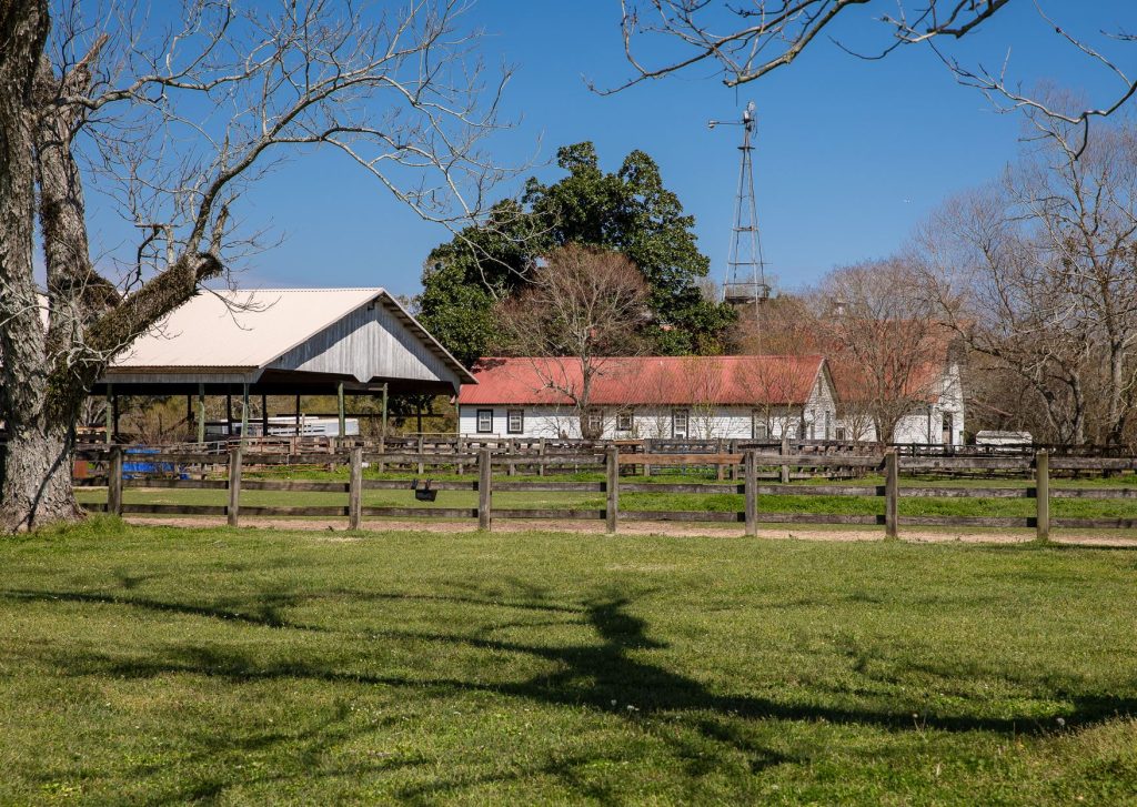 Wooden fence framing a pasture with a horse shelter, white farm buildings, and trees against a clear blue sky.