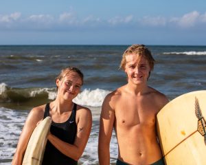 Two young surfers, a woman and a man, standing on the shore with surfboards, smiling after a surfing session, with waves in the background