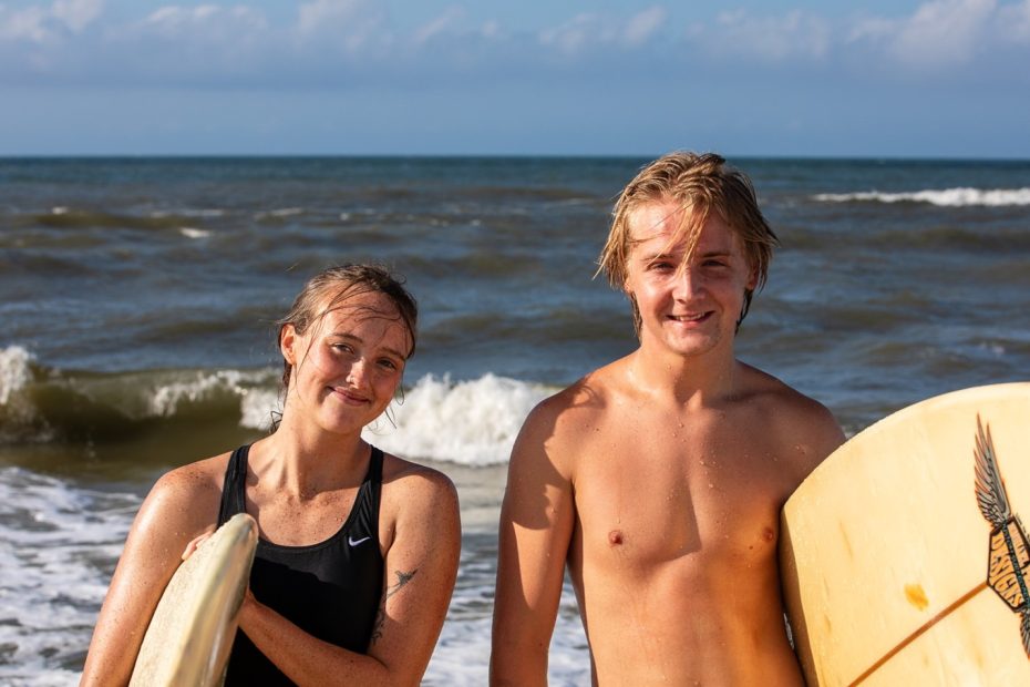 Two young surfers, a woman and a man, standing on the shore with surfboards, smiling after a surfing session, with waves in the background