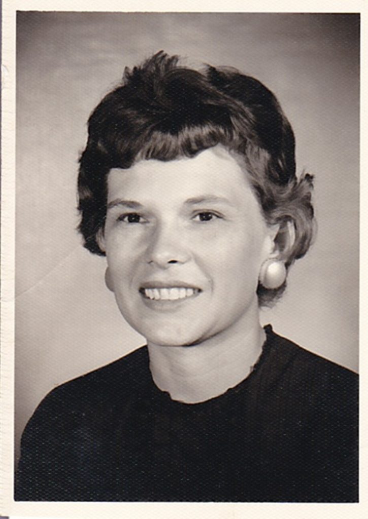 Vintage black and white portrait of a middle-aged woman smiling, dressed in a dark blouse with a round collar and pearl earrings, likely taken in the mid-20th century.