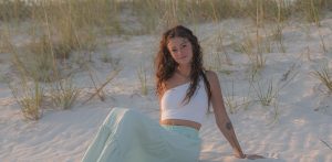 A young woman with curly hair, wearing a white crop top and a light green flowing skirt, is sitting on soft white sand at the beach. She is relaxed, leaning back slightly with her hand supporting her, surrounded by tall grasses in the background. The sun is setting, casting a warm, golden light over the scene.