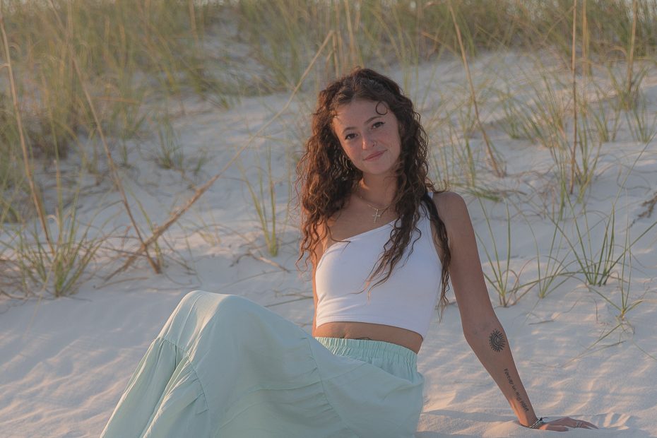 A young woman with curly hair, wearing a white crop top and a light green flowing skirt, is sitting on soft white sand at the beach. She is relaxed, leaning back slightly with her hand supporting her, surrounded by tall grasses in the background. The sun is setting, casting a warm, golden light over the scene.